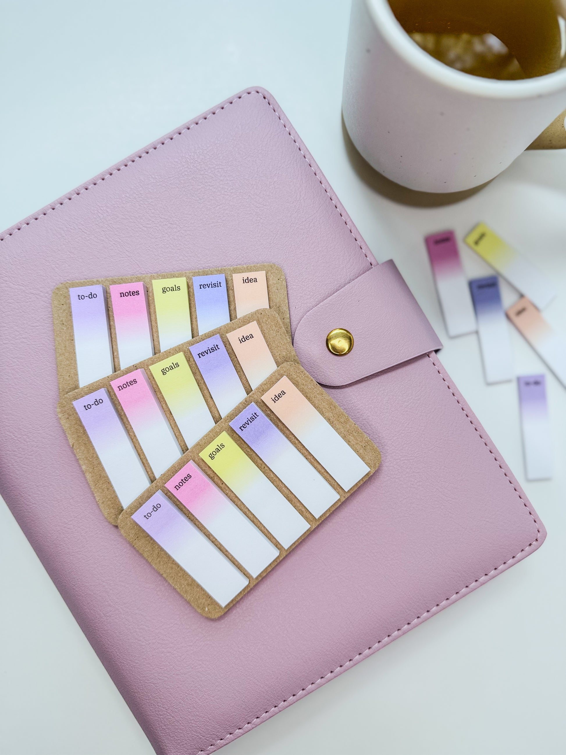 Pastel tabs for planners with the words to-do, notes, goals, revisit and idea next to a cup of tea and on top of a planner or agenda cover.