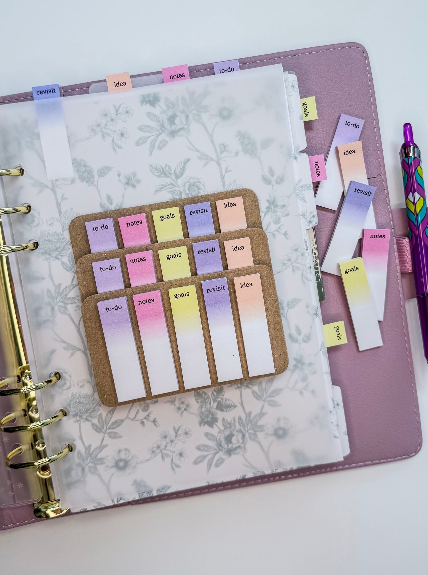 Pastel tabs for planners with the words to-do, notes, goals, revisit and idea on top of a planner and with a purple pen nearby. 